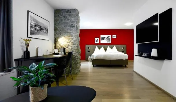 Open-plan room with stone accent wall, red accent wall, wall-mounted TV, desk with kettle and coffee maker, and bed with white linens at Hotel Zugertor in Zug