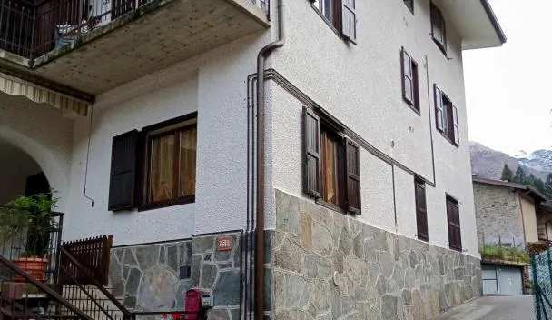 White two-story house with stone base, second-floor balcony, shuttered windows and gated entrance on a narrow street near mountains at Appartamento nonna Piera in Casargo