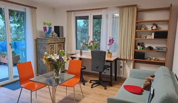 Open-plan living/dining with glass table and orange chairs, desk with ergonomic chair, sofa and garden access via sliding doors at Chalet Gold in Affoltern