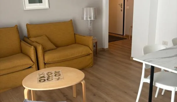 Open-plan living area with mustard sofa, round coffee table, lamp, basket and white dining table with two chairs at Verde Acqua Suite in Lecco