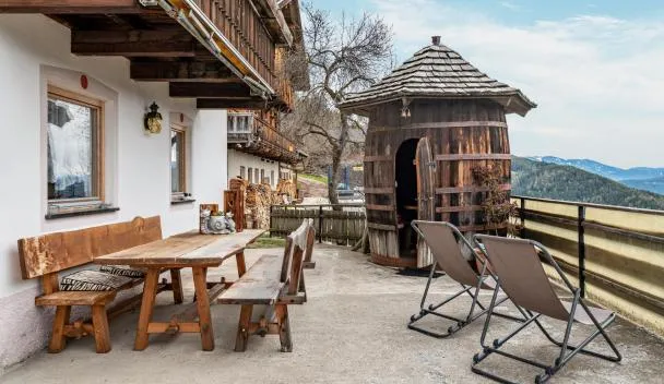 Outdoor terrace with wooden dining table and benches, two sun loungers and a wooden outhouse overlooking forested hills at Stöcklhuberhof in Vandoies