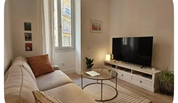 Living room with beige sofa, orange cushion, flat-screen TV on low console, round coffee table, woven rug and balcony window at 3 pièces - 2 chambres - Port de Nice in Nice