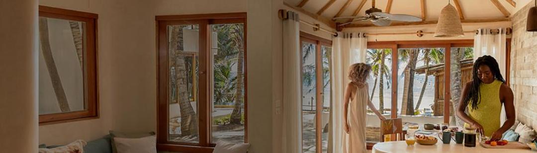 Una cocina luminosa y acogedora diseñada en piedra y madera natural con grandes puertas de cristal que dan a una playa tropical. En las ventanas hay cortinas finas que dejan pasar la luz dorada del sol. Dentro hay dos mujeres: una lleva un vestido vaporoso, está de pie junto a las puertas y está agarrando una silla. La otra lleva un top veraniego y está cortando fruta en la isla de la cocina. En la encimera hay una tetera, una piña y otros artículos de cocina. Detrás de las puertas, hay palmeras y una playa de arena que se extienden hasta el océano.