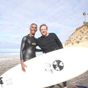 two men standing on the beach holding a surfboard