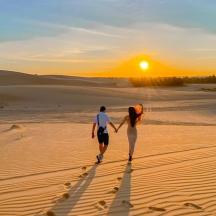 a man and woman walking through the desert at sunset