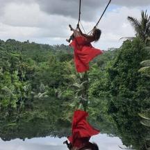 a girl in red dress swinging on a swing over a river
