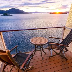 Sunset view from a cruise ship balcony with two empty wooden deck chairs and a small round table overlooking calm ocean water and distant forested islands.