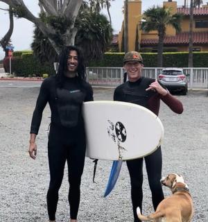两名身穿潜水服的女子拿着冲浪板和一只狗