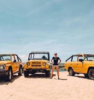 一名女子站在沙地上，旁边是两辆黄色卡车