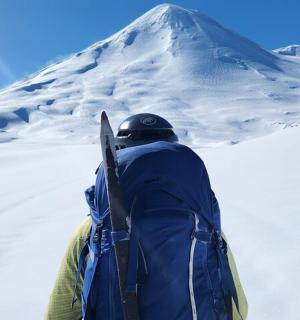 一名男子背着背包，带着滑雪板，站在白雪皑皑的山上
