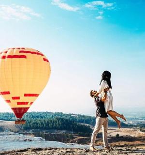 a man and woman standing on top of a hill with a hot air balloon