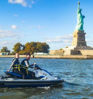 两个人骑着水上摩托艇在水面上，旁边是自由女神像。