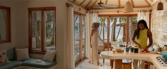 Una cocina luminosa y acogedora diseñada en piedra y madera natural con grandes puertas de cristal que dan a una playa tropical. En las ventanas hay cortinas finas que dejan pasar la luz dorada del sol. Dentro hay dos mujeres: una lleva un vestido vaporoso, está de pie junto a las puertas y está agarrando una silla. La otra lleva un top veraniego y está cortando fruta en la isla de la cocina. En la encimera hay una tetera, una piña y otros artículos de cocina. Detrás de las puertas, hay palmeras y una playa de arena que se extienden hasta el océano.