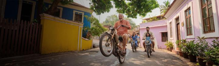 A smiling older man rides a motorcycle alongside his family, who are also riding motorcycles, on a tree-lined street.