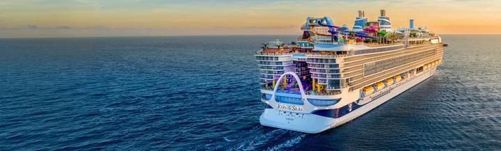 High-angle view of the Royal Caribbean cruise ship Icon of the Seas navigating calm blue ocean waters. Bright multicolored water slides visible on the top deck and rows of passenger balconies lining the side beneath a warm, golden horizon.