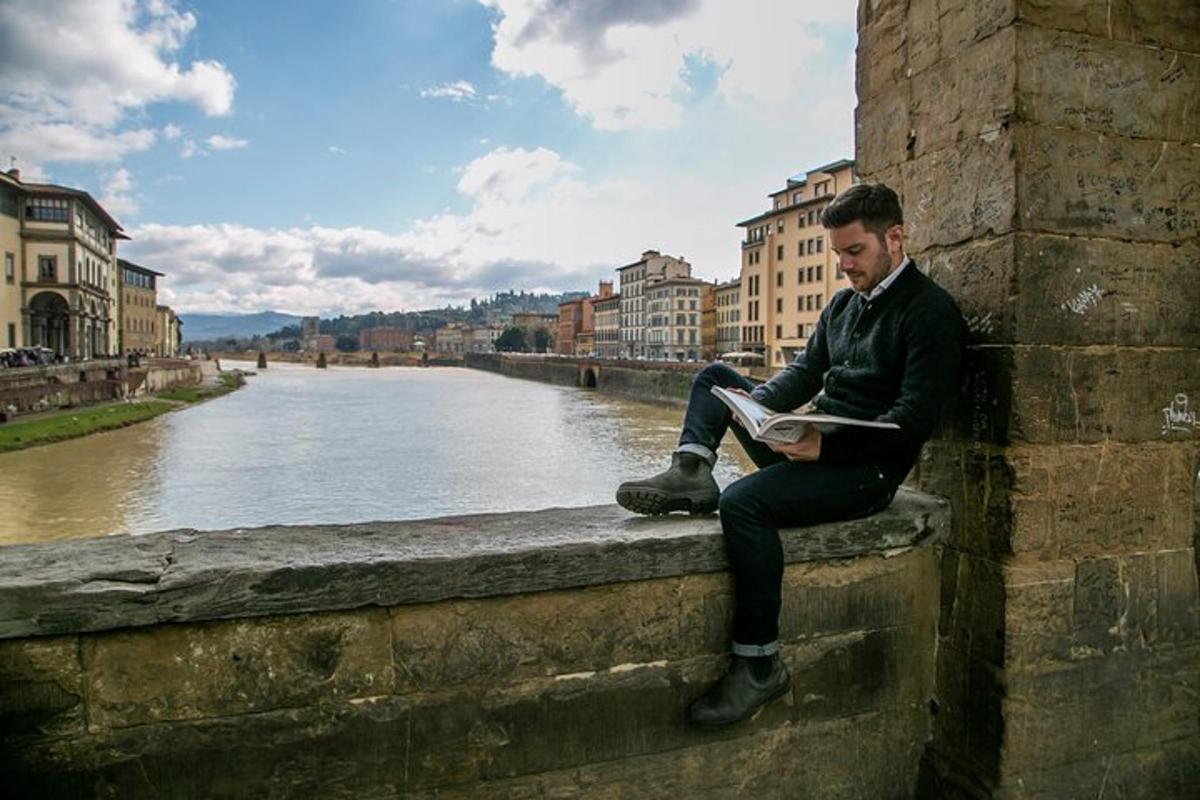 um homem sentado em um muro lendo um livro próximo a um rio