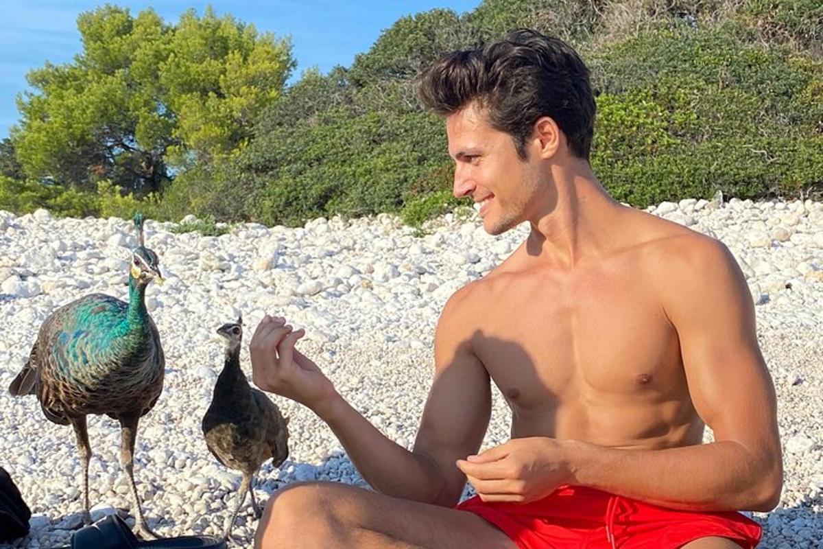 ein Mann ohne Hemd in roten Shorts sitzt neben einem Vogel