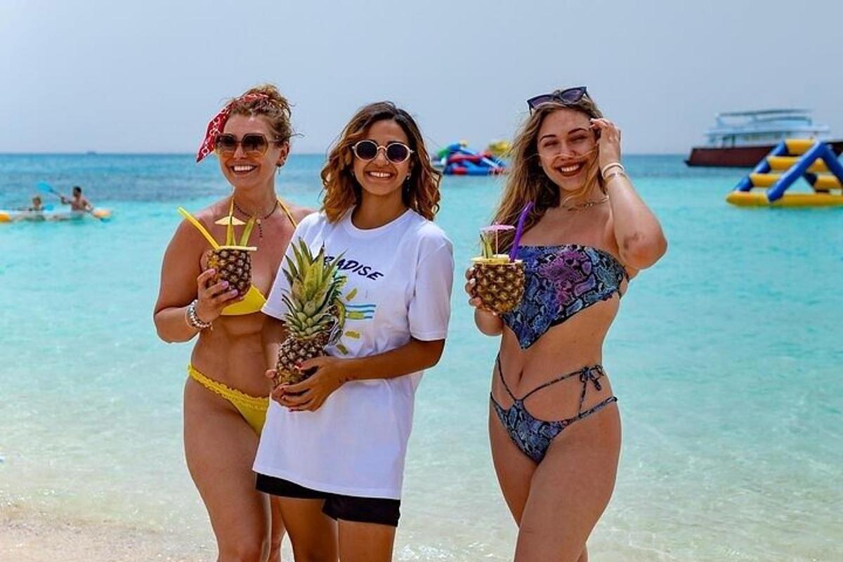 tiga wanita berdiri di pantai sambil memegang minuman nanas