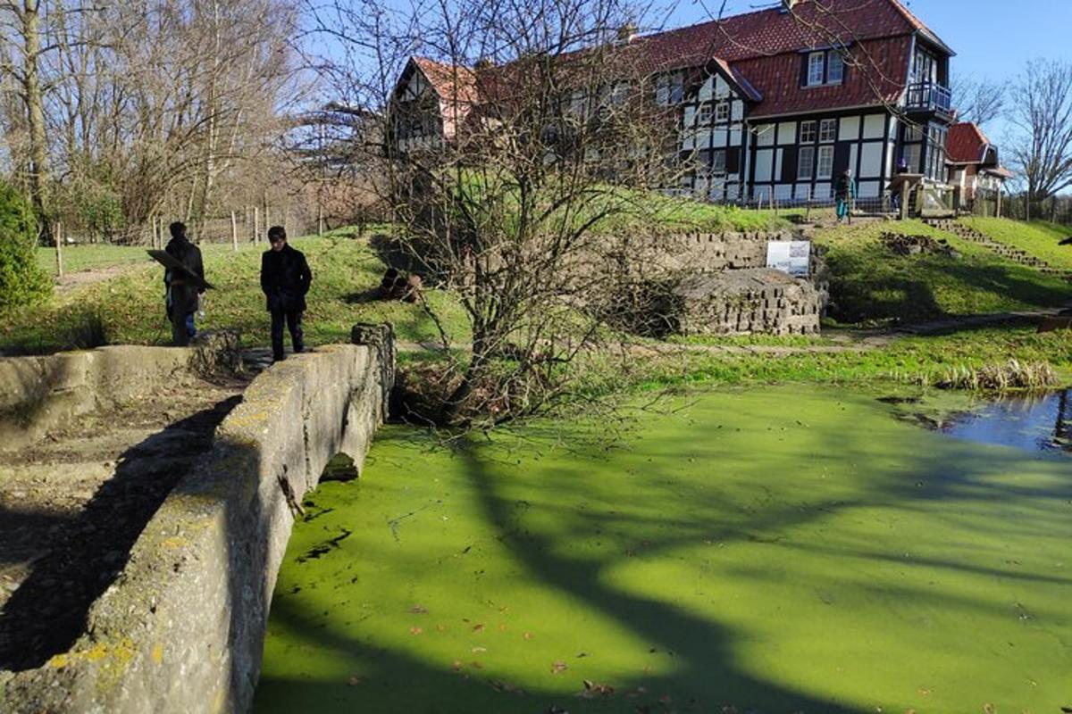 Dos personas caminando sobre un puente sobre agua verde frente a una casa.