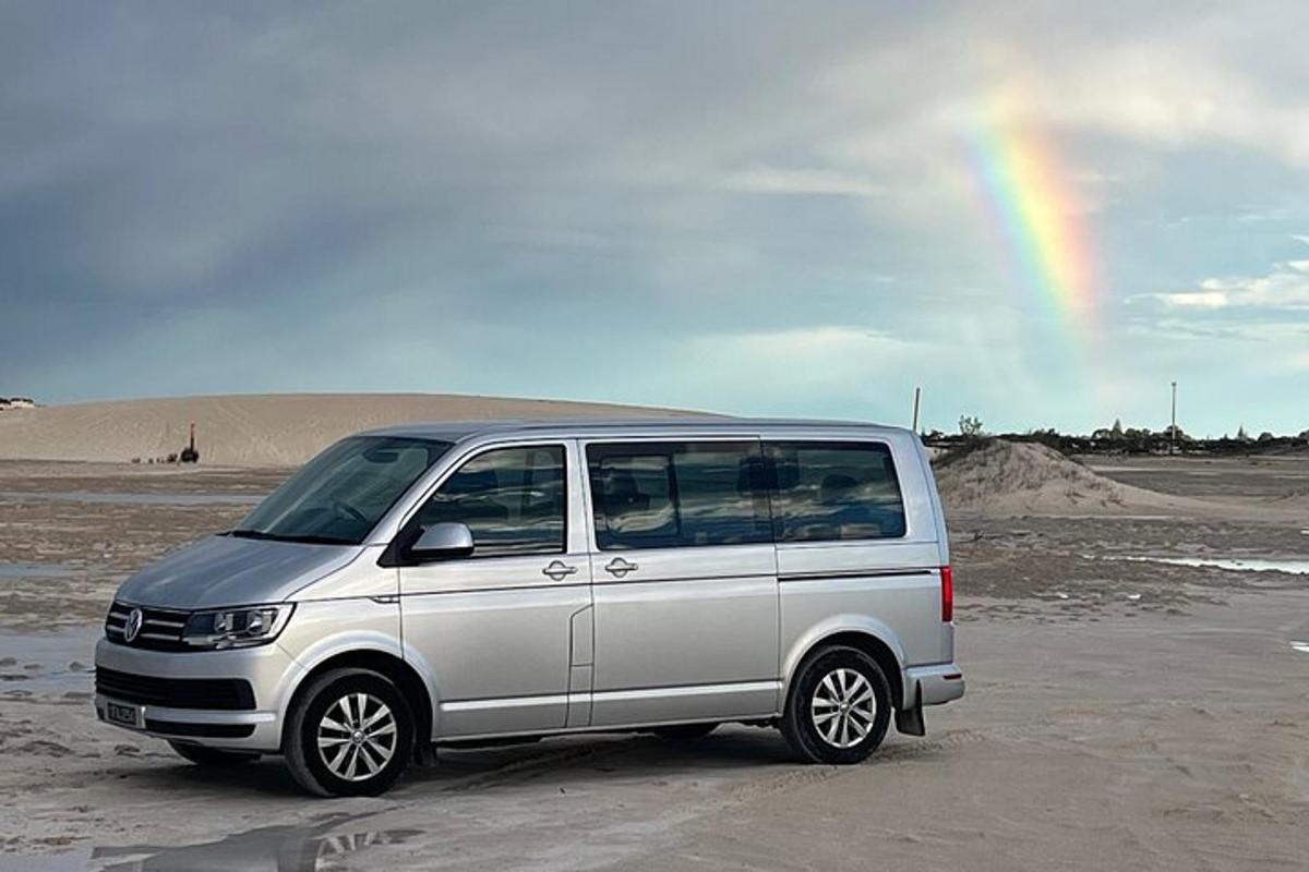 Una camioneta blanca estacionada en una playa con un arcoíris al fondo.