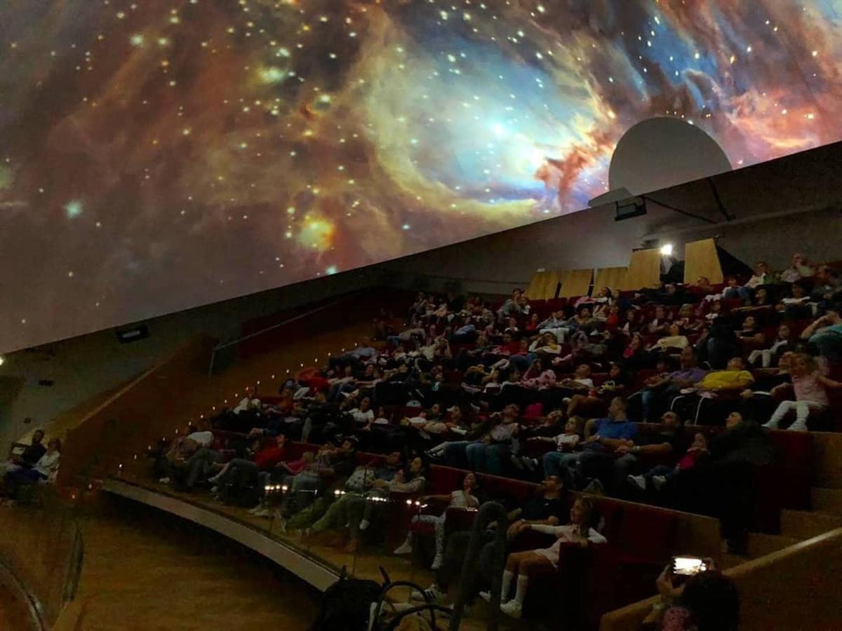 dav lidí sedících v hledišti s velkou nástěnnou malbou galaxie