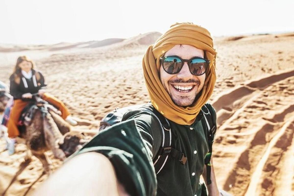 ein Mann mit Kopftuch und Sonnenbrille auf einem Kamel in der Wüste
