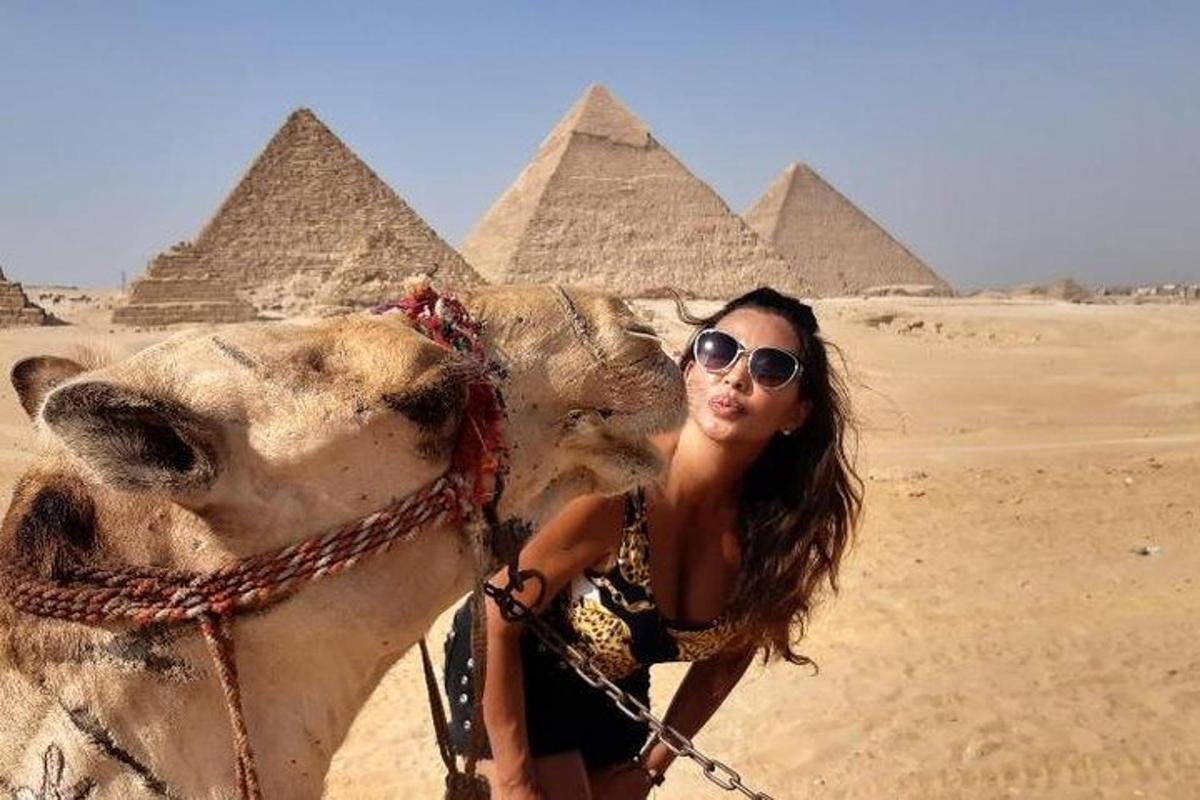 Una mujer parada junto a un camello frente a las pirámides.