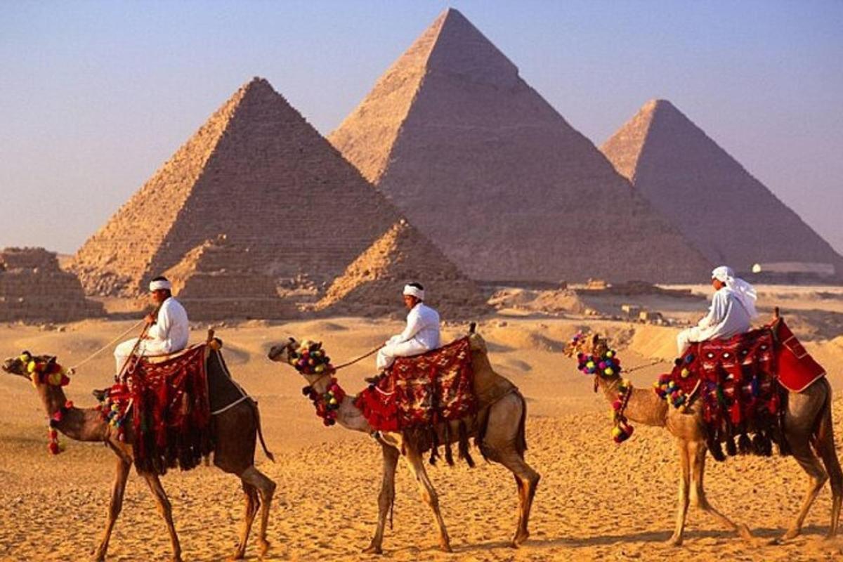 tři muži jedoucí na velbloudech před pyramidami