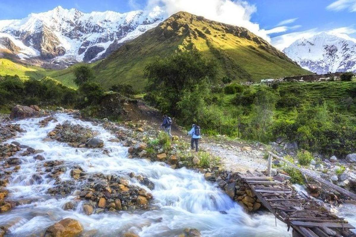 divi cilvēki stāv uz tilta pār upi ar kalniem