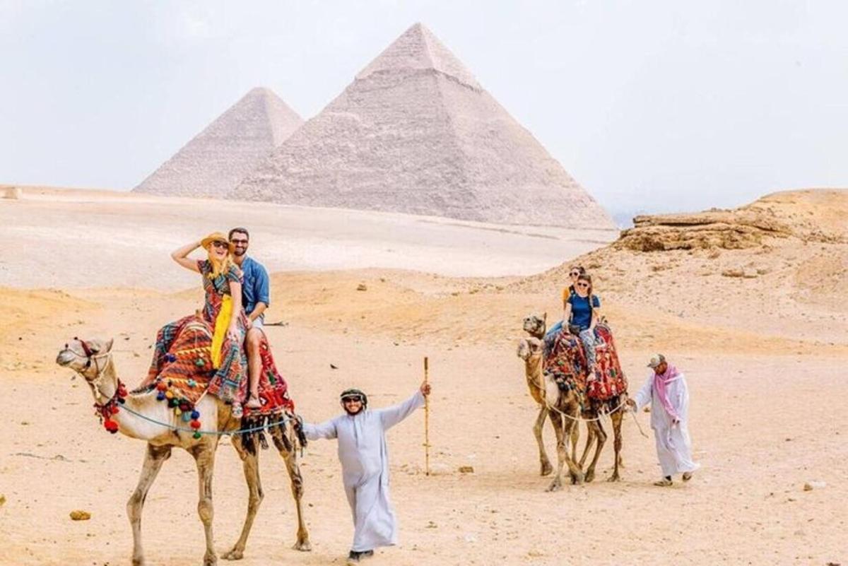 Skupina lidí jedoucích na velbloudech před pyramidami