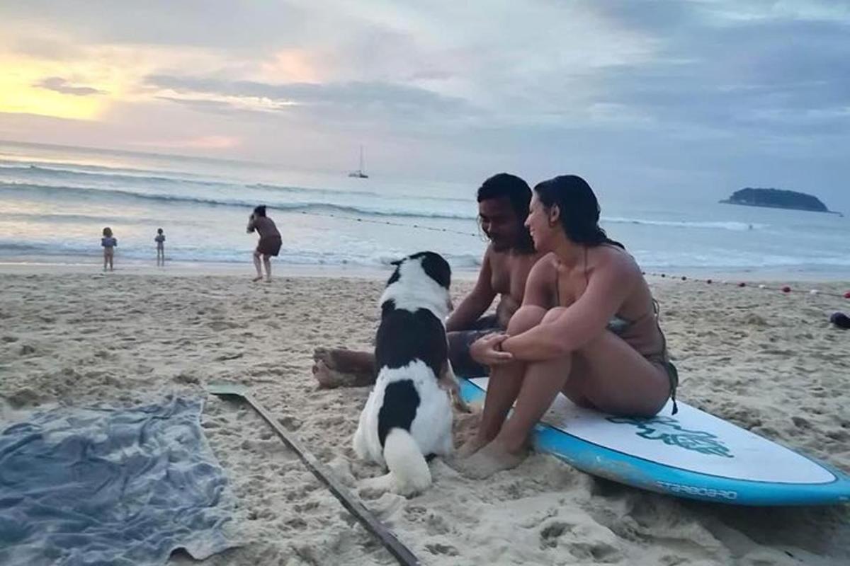 dues persones i un gos asseguts en una taula de surf a la platja