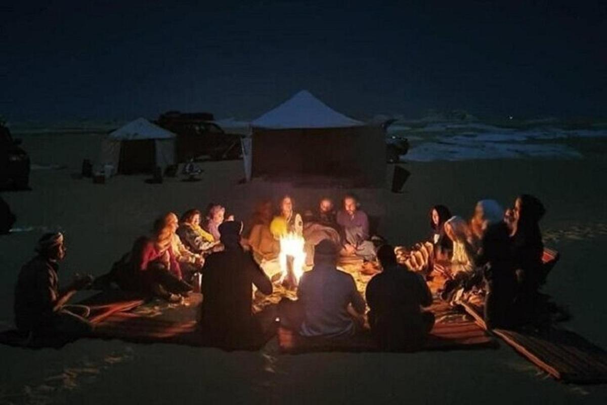 hópur fólks situr við eld á ströndinni að kvöldi