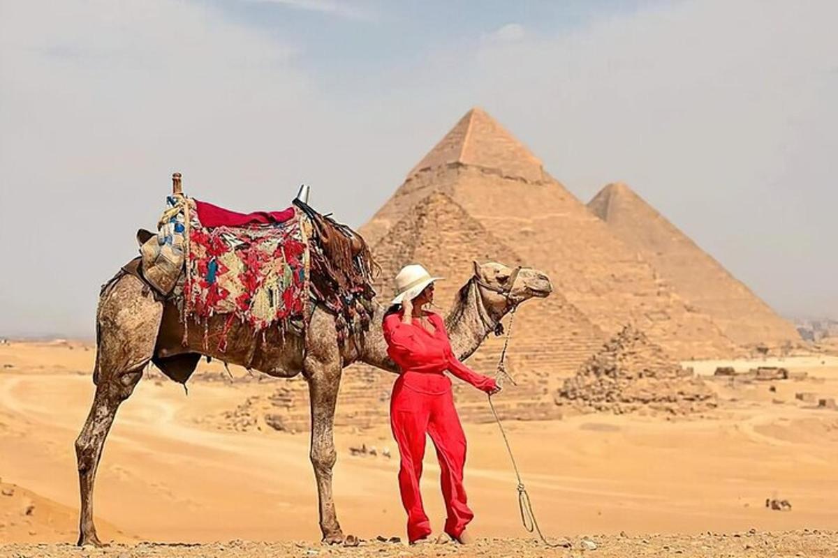 Una mujer parada junto a un camello frente a las pirámides.