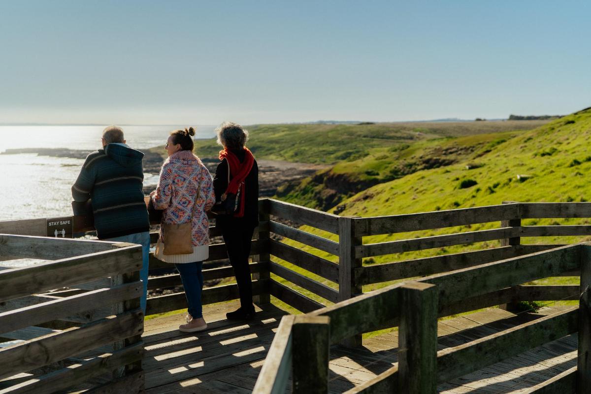 drei Personen stehen auf einem Holzsteg und blicken auf das Meer