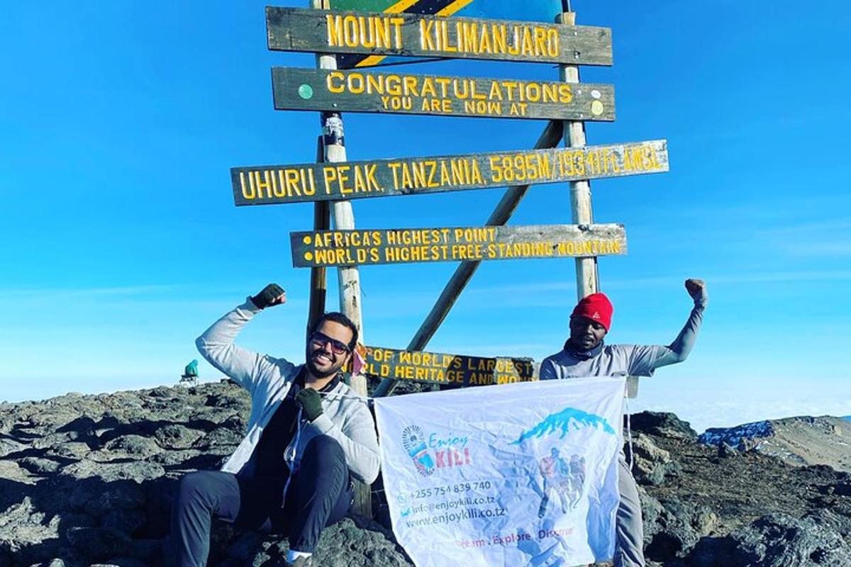 two men are posing in front of a sign on a mountain