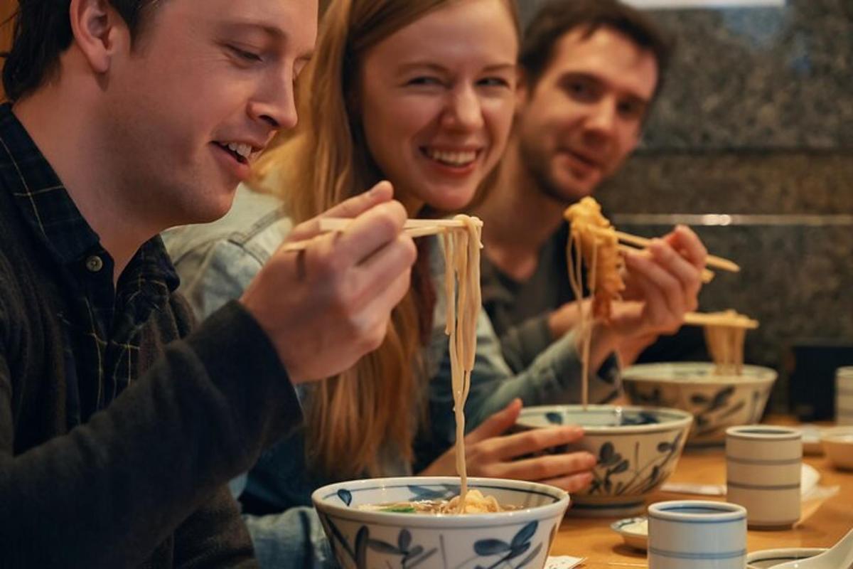 tři lidé sedí u stolu a jedí nudle hůlkami