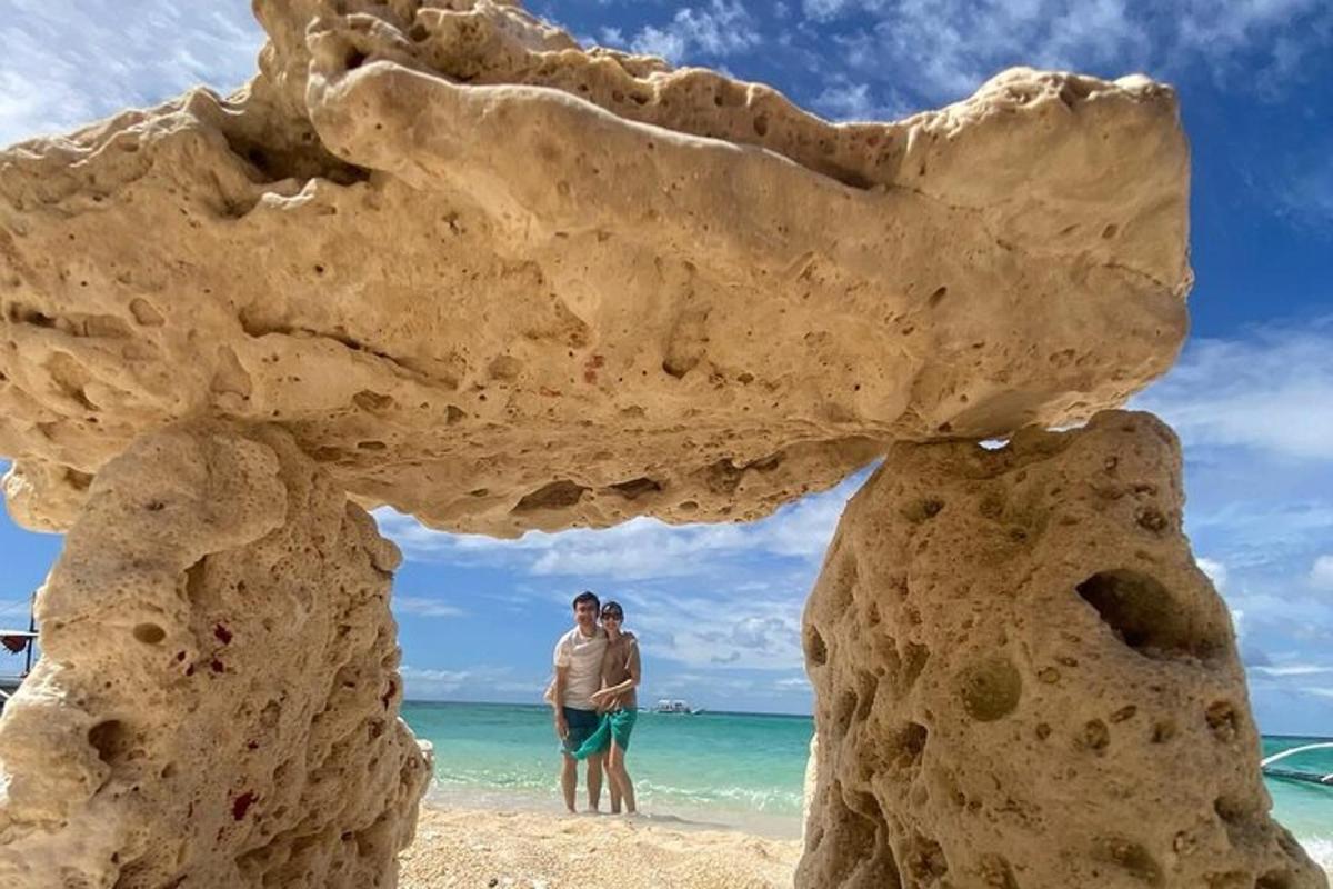 two people standing on the beach behind a rock formation