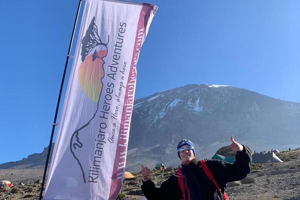 Un hombre de pie junto a una bandera con una montaña al fondo.