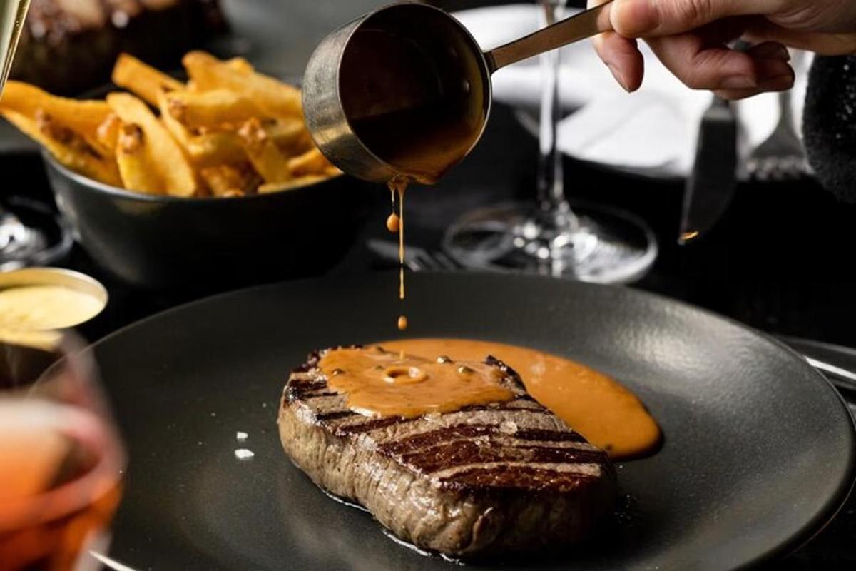 a person drizzling sauce on a steak with fries