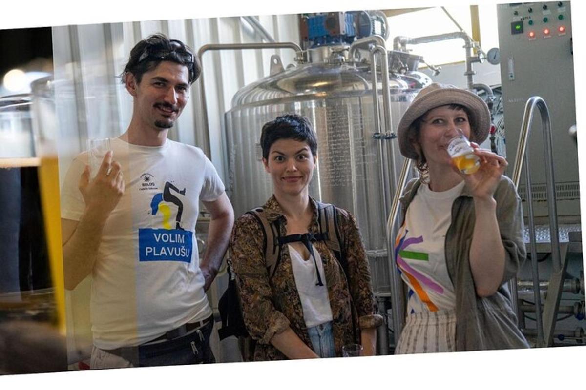 trīs cilvēki stāv alus darītavā ar vīrieti, kurš dzer no glāzes