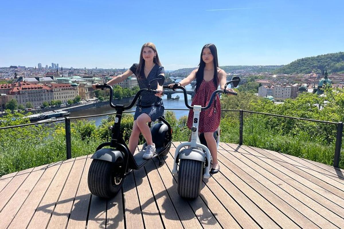 Két lány robogókon áll egy sétányon