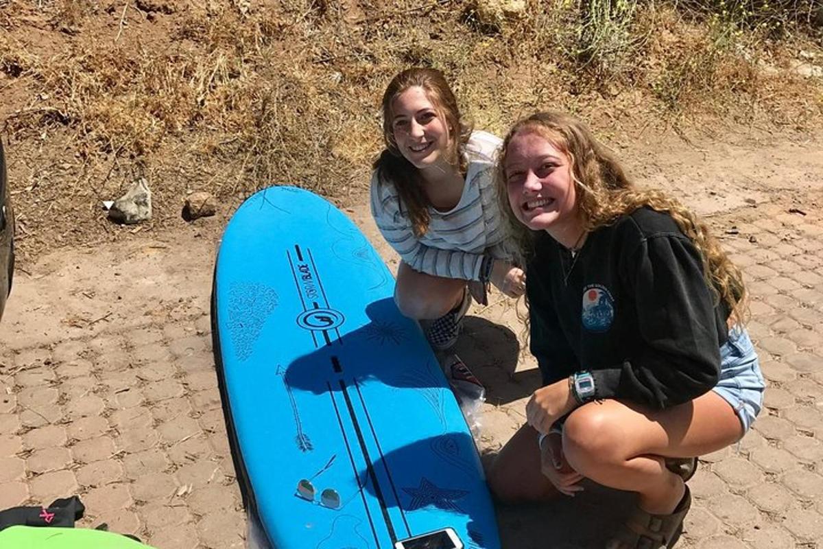 Dos mujeres están arrodilladas junto a una tabla de surf azul.