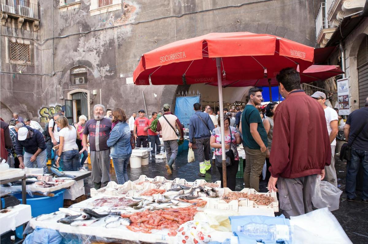 a group of people standing around a market with a red umbrella